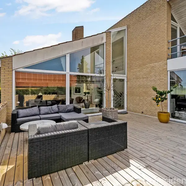 Sorte udemøbler står på en træterrasse foran et moderne hus med store glasvinduer. Terrassen er omgivet af planter i krukker, og himlen er klar med få skyer.