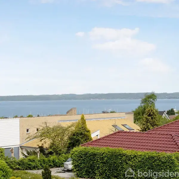 Rødt tegltag ses i forgrunden, omgivet af grønne buske. I baggrunden strækker en blå fjord sig ud mod horisonten under en klar, let skyet himmel.