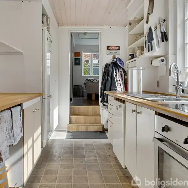 Et hvidt køkken med træbordplader har opvasken placeret ved vinduet. Knive hænger på væggen. Gangen fører til et andet rum i huset. Tøj hænger foran trappen.