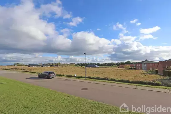 En bil holder på en stille vej ved siden af en græsklædt mark. I baggrunden ses røde bygninger under en blå himmel med skyer.