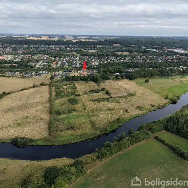 Landbrugsområde markeret med en rød pil i midten. En flod snor sig gennem landskabet. I baggrunden ses en by med spredte bygninger omgivet af marker og skove.