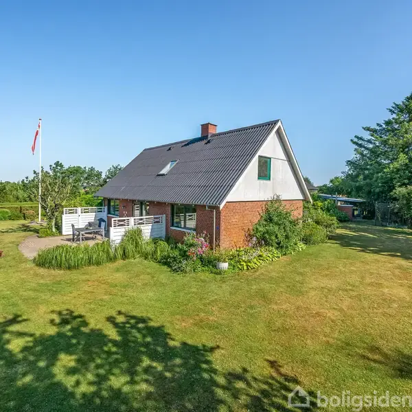 Et rødt murstenshus med gråt tag står i en velplejet have. En flagstang ses ved siden af huset. Omkring huset er der græsplæne og grønne træer under en blå himmel.