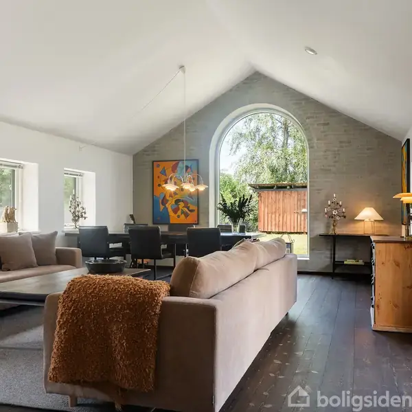 En lys stue med to beige sofaer, et spisebord og stole. Store vinduer og kunst på væggene skaber en moderne atmosfære. Et tæppe ligger på det mørke gulv.