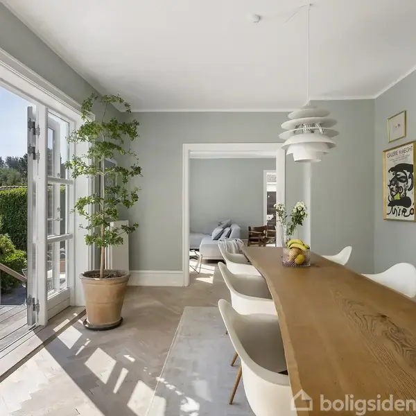 Spisebordet er omgivet af hvide stole i et lyst rum med åbne døre til en træterrasse. Væggene er dekoreret med kunst, og en stor plante står ved døren.