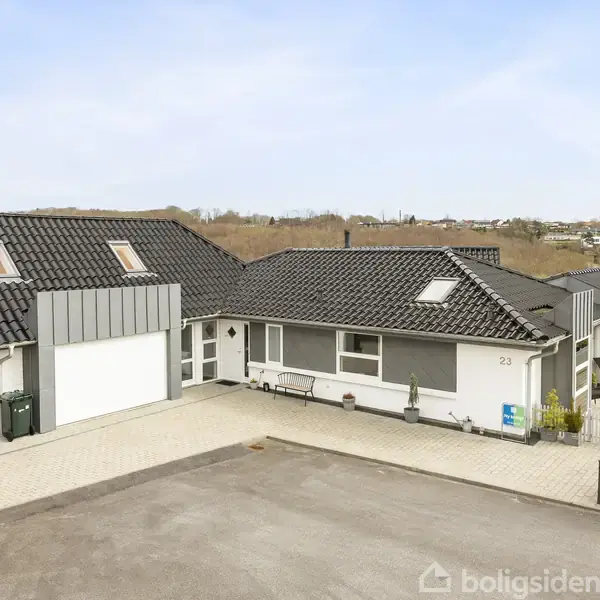 Et moderne hus står på en flisebelagt indkørsel med gråt tag og hvide vægge. Omkring huset er der en velholdt have og buske. Nummeret "23" ses på væggen.