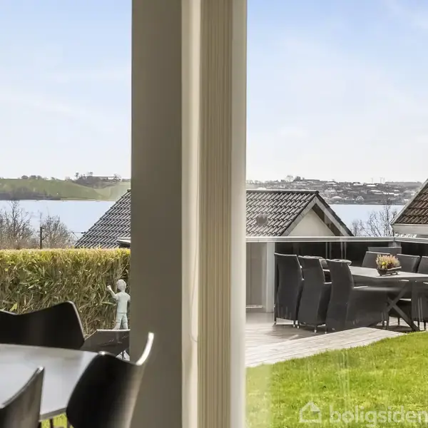 Et bord står i forgrunden ved siden af store vinduer. Udendørs ses havemøbler på en terrasse, omgivet af grønne buske, med en udsigt over vand og huse i det fjerne.