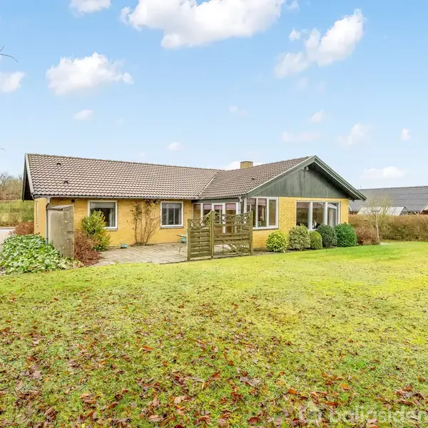 Et gult, én-etagers hus står stille i en have med en velplejet græsplæne. Huset har en tagterrasse, omgivet af buske og trækonstruktioner på en lys dag.