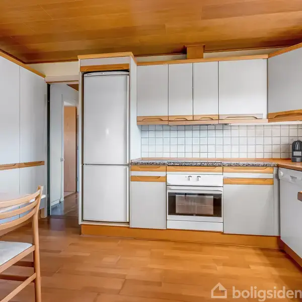 Et lyst køkken med hvide skabe og trædetaljer. Køleskab, ovn og opvaskemaskine er integreret. Et bord med to stole står til venstre, omgivet af glatte trægulve og lyse vægge.