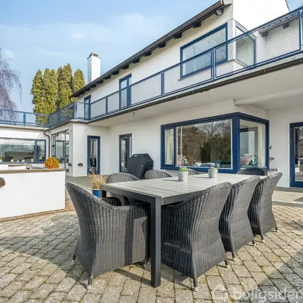 En udendørs terrasse med et langt spisebord og seks flettede stole, beliggende foran et moderne to-etagers hus med store vinduer og blå detaljer, omgivet af træer.