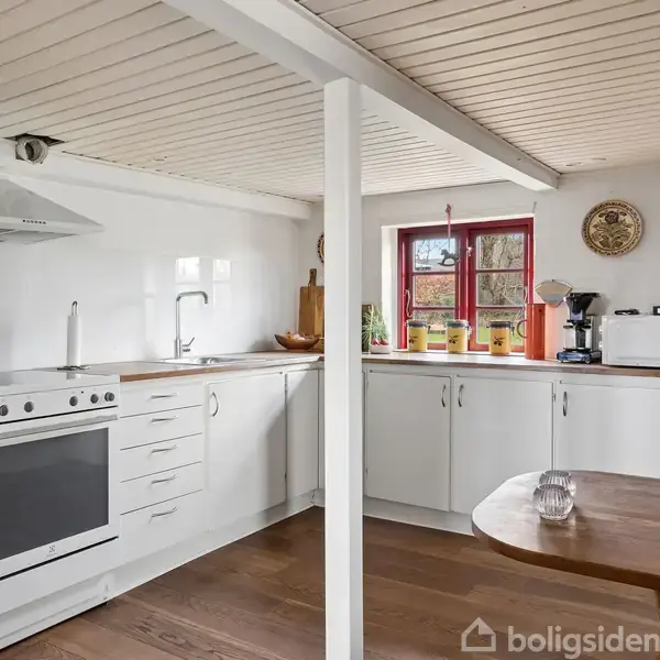 Køkken med hvide skabe og en indbygningsovn under en emhætte. Et bord med glas står ved siden af. Vindue og kaffemaskine er i baggrunden. Ingen tekst.