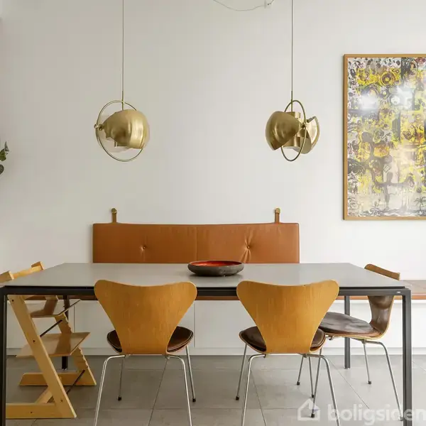 En spisebordsopsætning med fire moderne træstole omkring et bord, en brun læderbænk bagved. To gyldne loftlamper hænger over bordet. En farverig kunstplakat hænger på væggen.
