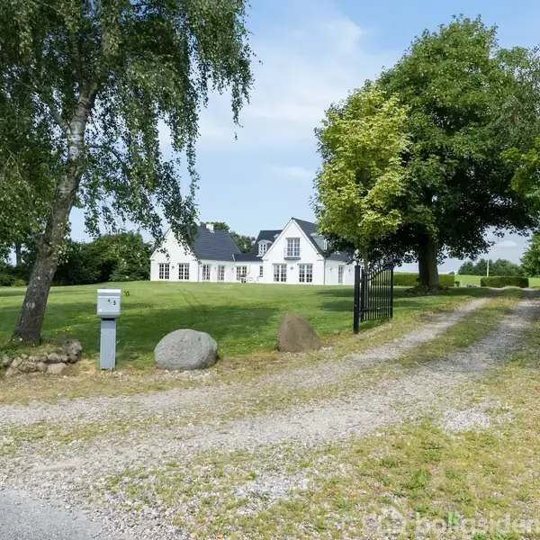 Hvid hus med sort tag står i bunden af en grusvej omgivet af grønne træer og græsplæne. Sten og postkasse er ved vejen.