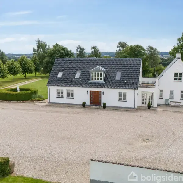 Hvidt, moderne hus med sort tag står på en stor grusindkørsel. Omkringliggende grønne marker og træer skaber en landlig atmosfære under en klar blå himmel.