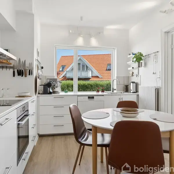 Et hvidt, moderne køkken med en rund spisebord omgivet af stole; i baggrunden ses en stor rude med udsigt til et hus med rødt tag samt flere køkkenredskaber og -apparater.
