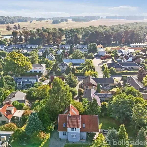 Huse ligger tæt i en landsby omringet af frodig vegetation. Røde tegltage er synlige fra luften, mens marker og skove strækker sig i baggrunden under en klar himmel.