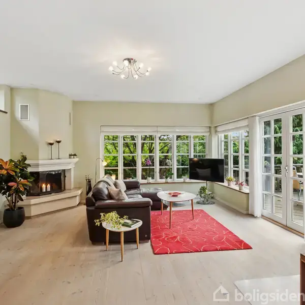 Stue med en sort sofa og rødt tæppe, udstyret med kamin, ved siden af store vinduer og en dør til en terrasse. Grønne planter og vægmaleri tilføjer farve.