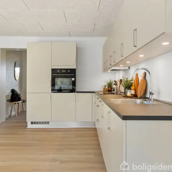 Et moderne køkken med beige skabe langs væggen. Indbygget ovn i højre side. Bordpladen er dekoreret med planter og skærebræt. Stue med lys træparketgulv ses i baggrunden.