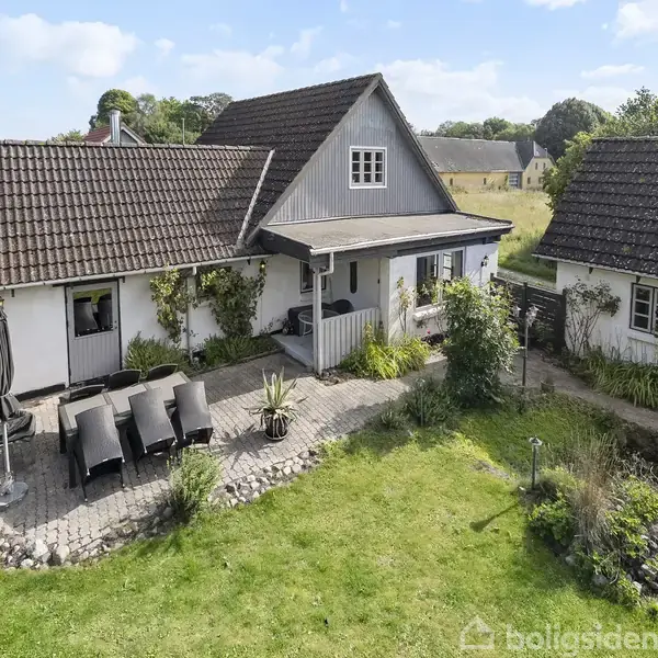 Hvidt hus med mørkt tegltag står i en frodig have. Foran terrassen står havemøbler og parasol. Baggrunden viser åbent, grønt landskab med enkelte træer og blå himmel.