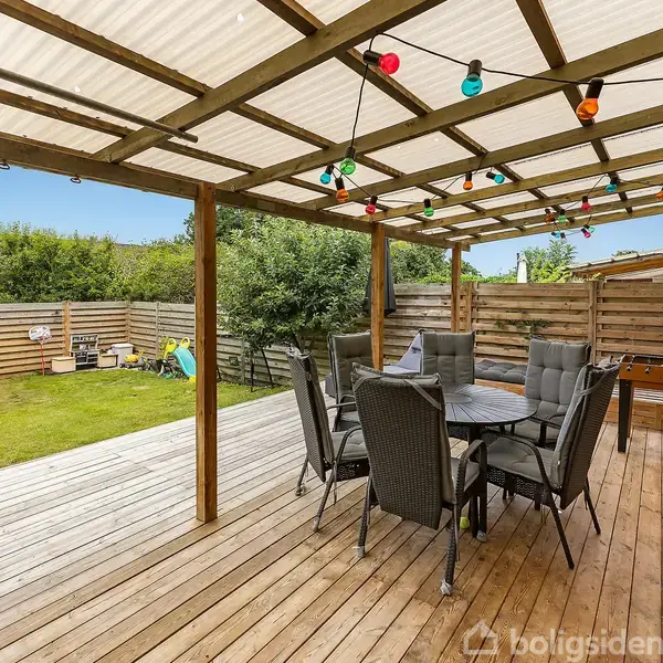 En overdækket terrasse med et rundt bord og stole. Farvede lyskæder hænger fra loftet. Ved siden af terrassen er der en græsplæne med legetøj og indhegning.