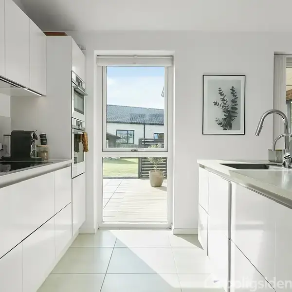 Et moderne køkken med hvide skabsfacader og indbygget ovn. En kværn står på bordet, og store vinduer slipper sollys ind fra terrassen. Et billede hænger på væggen.