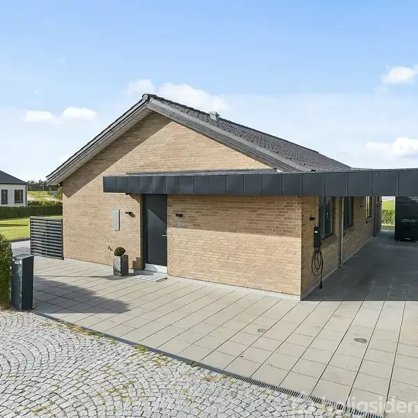 En enkelt etagers murstensbygning står stille i et boligområde. Den har et sort tag, carport ved siden og brostensbelagt indkørsel. I baggrunden ses en anden lignende bolig.