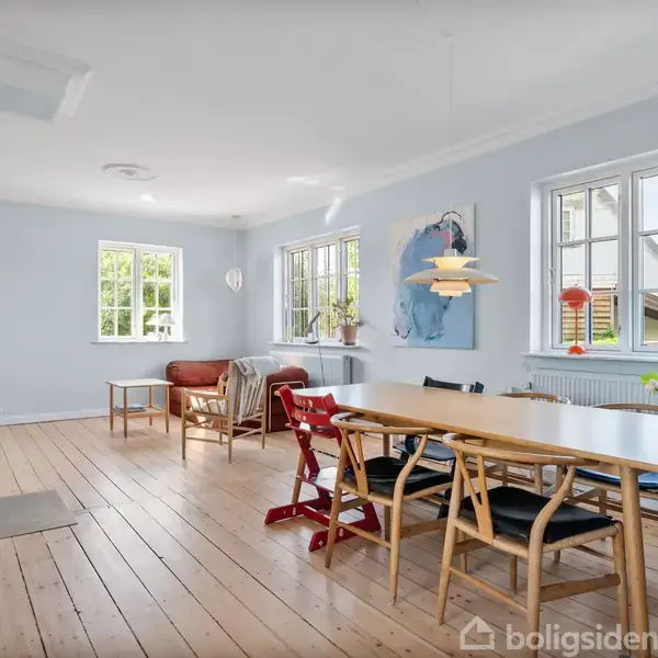 Spisebord står med stole i et lyst, rummeligt rum. En brændeovn står til venstre, mens vinduerne giver udsigt til en grøn have. Et maleri hænger på væggen.