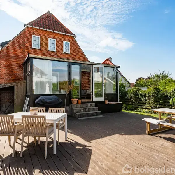 Hus i mursten med rødt tegltag har en stor træterrasse med spisebord og bænke. Glasudestue forbinder huset til terrassen. Haven omkranser terrassen med grønne buske og hegn.