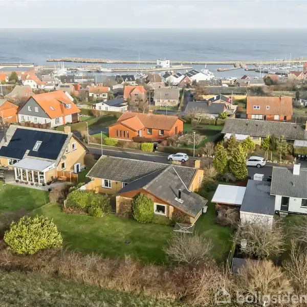 Flere huse ligger tæt sammen i en by nær kysten. Havenes grønne græs og buske omkranser husene, mens en havn og havet ses i baggrunden.