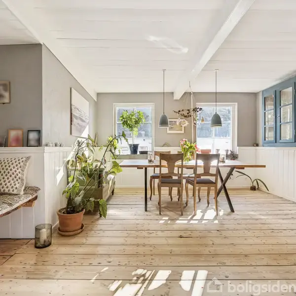 Spisebord står i en lys stue med trægulv og hvide vægge. Bordet er dekoreret med planter. Store vinduer slipper sollys ind, og der er en indbygget bænk med puder.