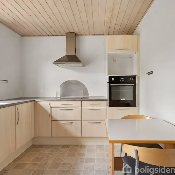 Køkken med lys træindretning, bordkomfur og indbygningsovn. Spisebord med to stole står ved siden af. Vinduet lader dagslys komme ind, og loftet er af træpaneler.