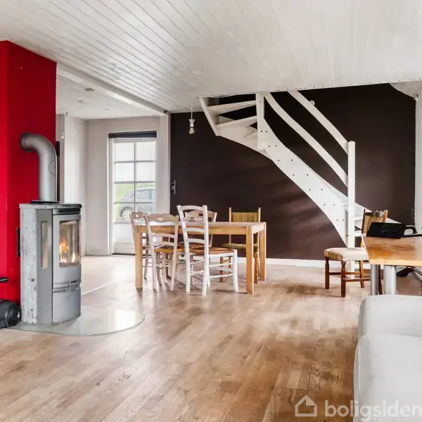 En brændeovn står i et åbent rum, omgivet af hvide vægge og en mørk accentvæg. Der er et spisebord med stole, en grå sofa, en støvsuger og en trappe.