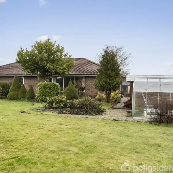 Murstenshus med rødt tag står i en frodig have med grønne buske og et drivhus ved siden af, omgivet af en velplejet græsplæne under en blå himmel.