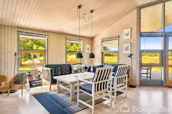 En stue med sort lædersofa og hvidt spisebord; på bordet er der en lampe over. Rummet har store vinduer med udsigt til en grøn have.