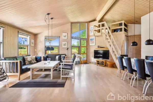 Moderne stue med lyst træ-interiør, sofa og spisebord, fjernsyn under en loftstue via trappe. Store vinduer giver udsigt til det grønne udenfor. To loftslamper hænger over bordet.