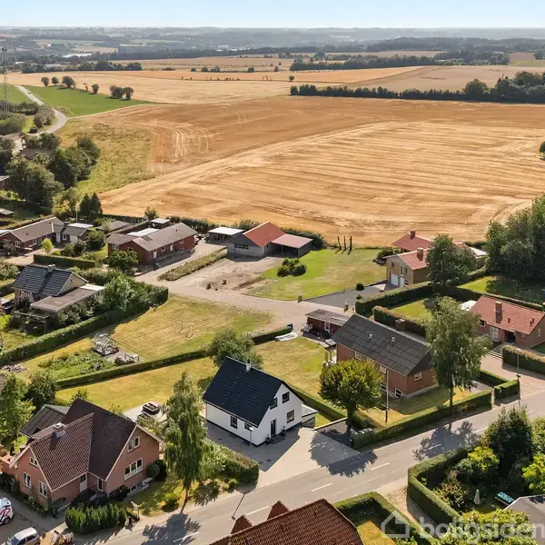 Bebyggelse med huse og haver omkranset af veje, beliggende ved siden af store gule marker under en tom himmel. Træer og buske bidrager til det grønne landskabsbillede.