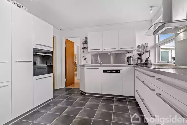 Et moderne køkken med hvide skabe, en indbygget ovn og en opvaskemaskine. Der er sort flisegulv, og køkkenet ligger i et lyst rum med vindue.