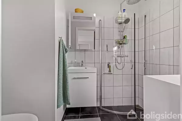 En moderne badeværelsesvask med skab nedenunder står foran et spejl. Ved siden af er der en brusekabine med glasdør. Håndklæde hænger på væggen. Gulvet er flisebelagt.