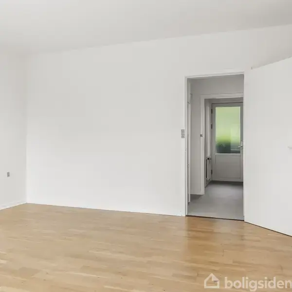 Tomt værelse med trægulv, åben dør mod korridor, der fører til et andet rum med et vindue. Lyse vægge og et enkelt, minimalistisk interiør.