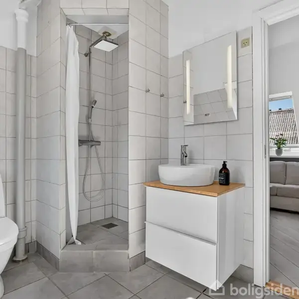 Toilet og bruser står i et flisebeklædt badeværelse. En håndvask med spejl hænger ved siden af. Gennem en åben dør ses en stue med sofa.