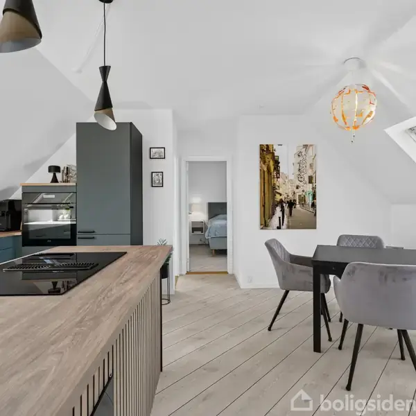 Et moderne køkken-alrum med kogeplade på en træbordplade, grå køkkenskabe, og en spisebord med fire stole under et loft med skråvinduer. Ingen tekst identificeret.