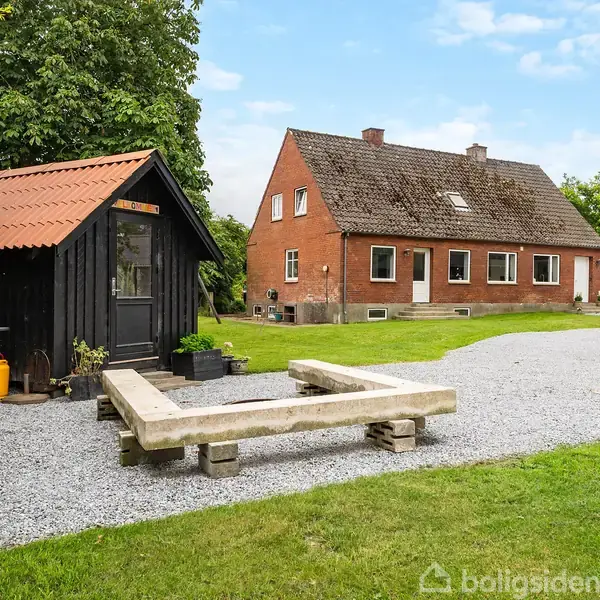 Et rødt murstenshus står i en grøn have med en grusbelagt sti. Et sort skur med rødt tag er placeret ved siden af en grill og en bænkeramme.