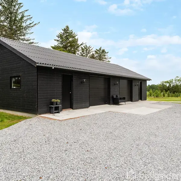 Sort, træbeklædt hus står på en gruset plæne. Ved siden af er en lille terrasse med stole. Omgivet af grønt græs og spredte træer under en klar himmel.