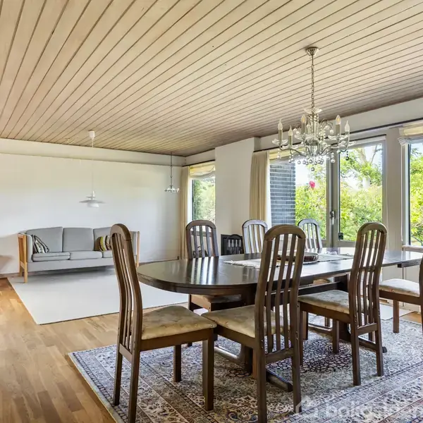 Spisebord med otte stole står på et tæppe i en stue. Store vinduer giver udsigt til en have. I baggrunden ses en sofa og lænestol.