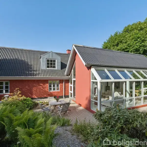 Rødt hus med sort tag, har et glashus ved siden af. Står i frodig have med terrasse, omgivet af grønne træer og blå himmel.