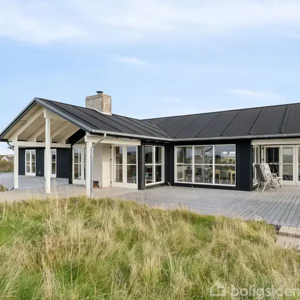 Sort hus med grå tag, der står i grønne omgivelser med grusvej foran. Flere huse er synlige i baggrunden under en blå himmel.