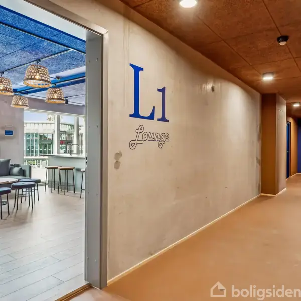 Dør fører til loungeområde med blå tekst "L1 Lounge" på en væg. Indenfor ses sofamøbler og bord i lys, moderne indretning under pendellamper.