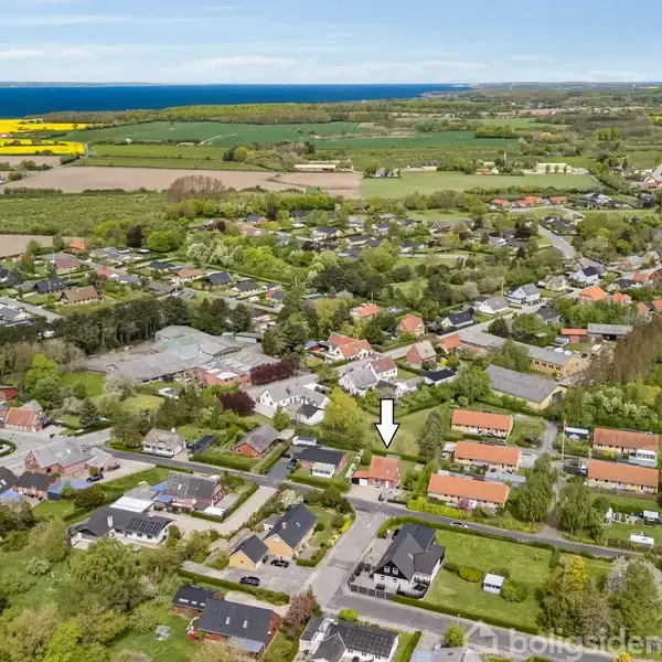 Pil peger på et hus i en boligområde med grønne omgivelser, tætliggende huse og marker i horisonten. Kystlinje og hav ses i baggrunden.