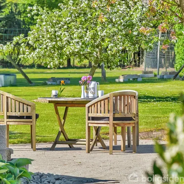 Et rundt træbord med to stole står på en terrasse omgivet af frodig have, blomster og grønne træer i baggrunden. En vase med blomster er placeret på bordet.