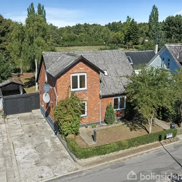Et rødt murstenshus med et gråt tag står langs en vej. Foran huset er en lille have med træer og buske. Huset har en satellitdisk monteret på siden.