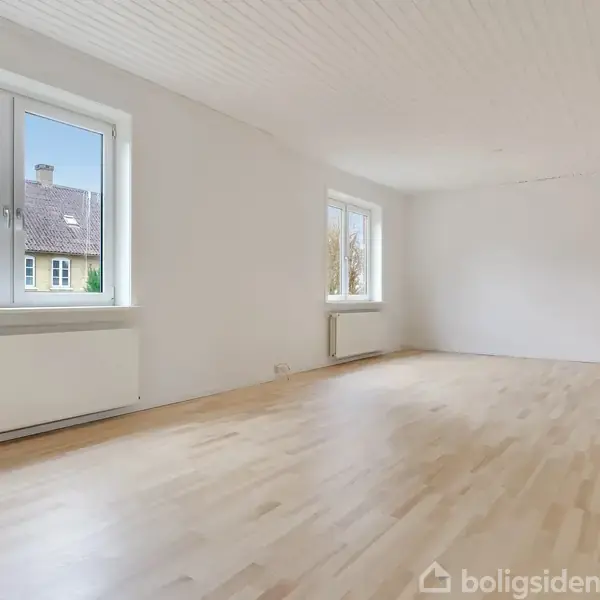 Et tomt, lyst værelse med trægulv og hvide vægge. To store vinduer giver udsigt til et rødt hus. Radiatorer under vinduerne.